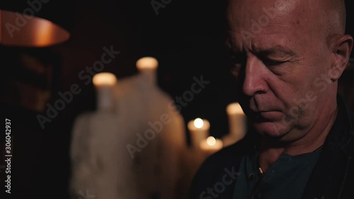 An elderly man is reflecting upon something. He is looking down and blinking in a half-light of many candles. A great art and social concept.
