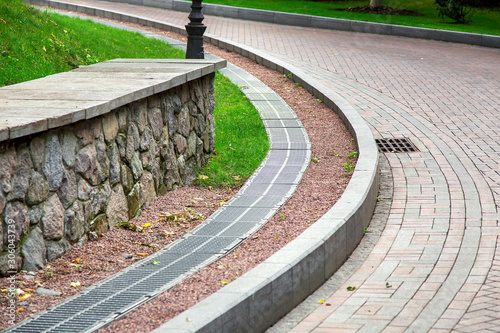 Φωτογραφία grille of the drainage system manhole on the curve pedestrian sidewalk in a turn on the rise up, on the side of the sidewalk a storm channel with an iron grate on the park