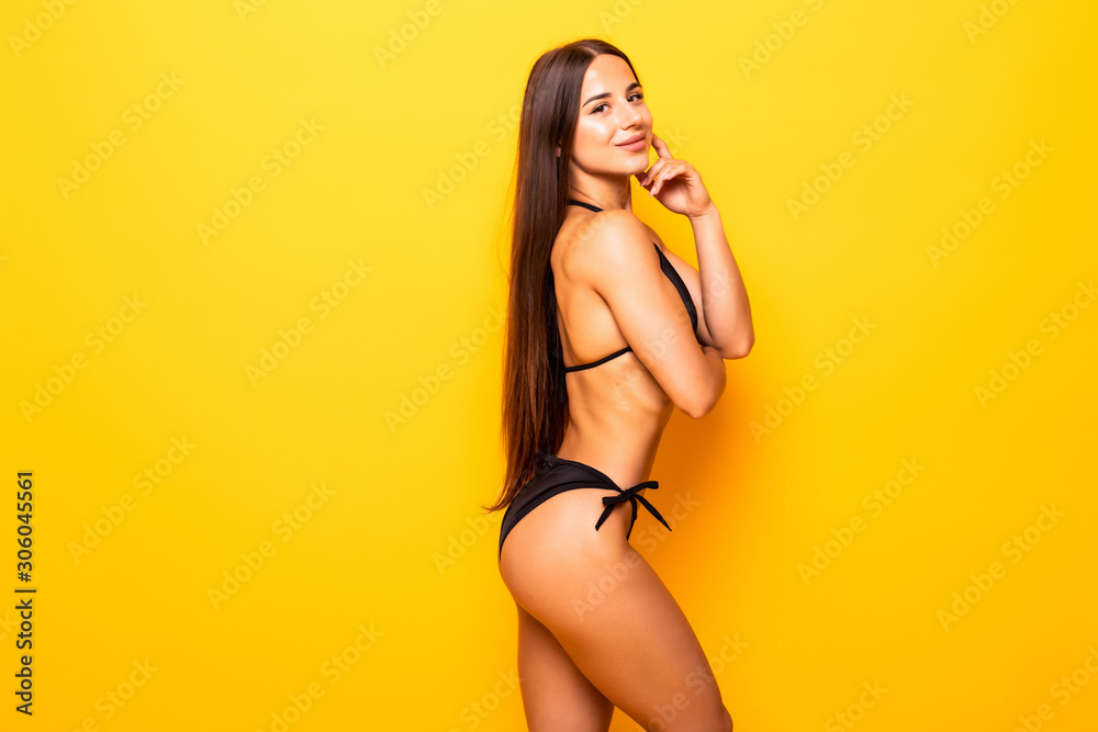 Young sexy slim tanned woman in black swimsuit posing against yellow background. - obrazy, fototapety, plakaty 