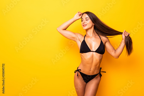 Smiling young woman posing with her black bikini swimsuit on yellow background