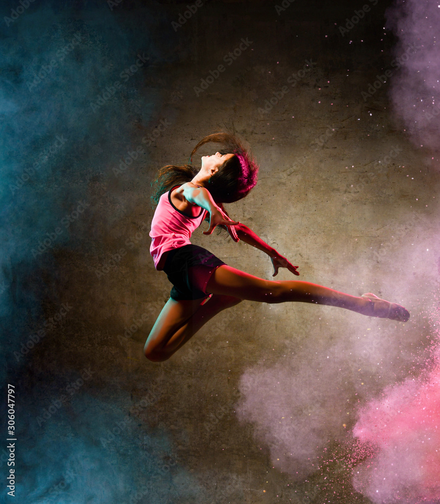 Street dance girl dancer jumping up dancing in neon light doing gymnastic exercises in studio on dark wall