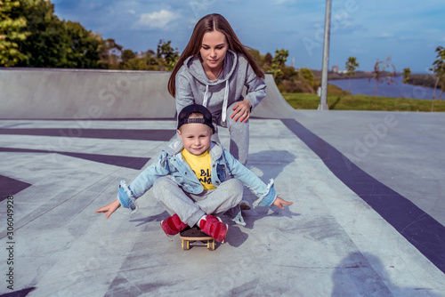 Young mother with little boy son, playground family learns ride skateboard, summer outdoors, autumn day. Emotions of delight, joy of happiness fun. Caring support assistance in training, parenting. © byswat