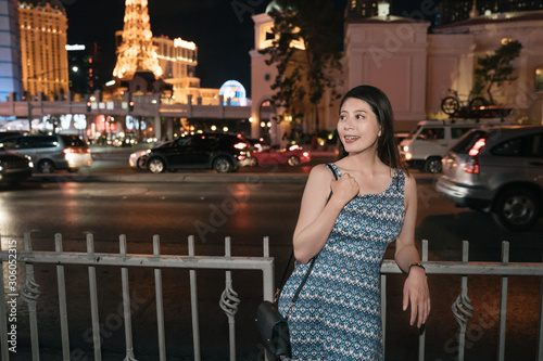 female traveler standing near road with car driving through and enjoying view and city lights in summer night in las vegas. young girl tourist leaning on railing and smiling relaxing on street.