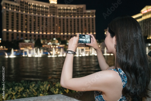 side view of beautiful young asian girl traveler watching fountain show and making photo on cellphone. elegant lady tourist using mobile phone taking picture with beautiful water dance at night