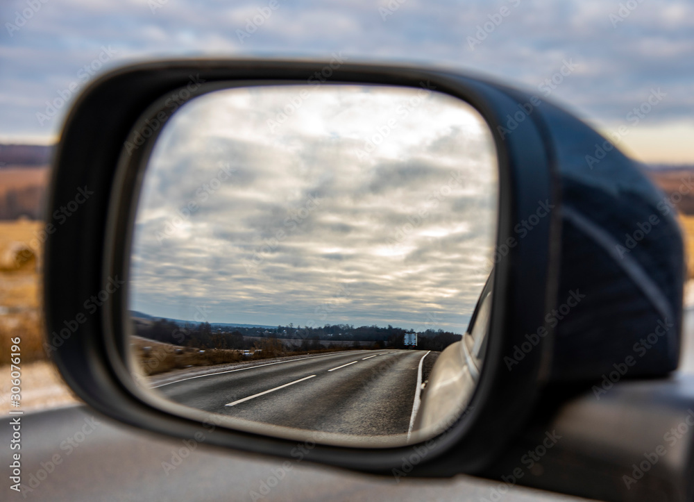 The road and the cloudy sky are reflected in the mirror of the car.