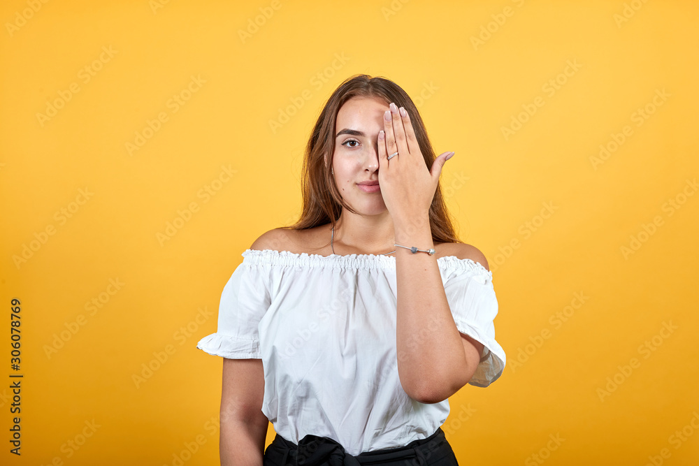 Charming caucasian brunette woman covering eyes with hand, looking serious isolated on orange background in studio in casual white shirt. People sincere emotions, lifestyle concept. Charming caucasian brunette woman covering eyes with hand, looking serious isolated on orange background in studio in casual white shirt. People sincere emotions, lifestyle concept.