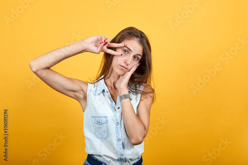 Boring caucasian young woman in blue denim shirt doing victory gesture, keeping hand on cheek, sleep isolated on orange background in studio. People sincere emotions, lifestyle concept.