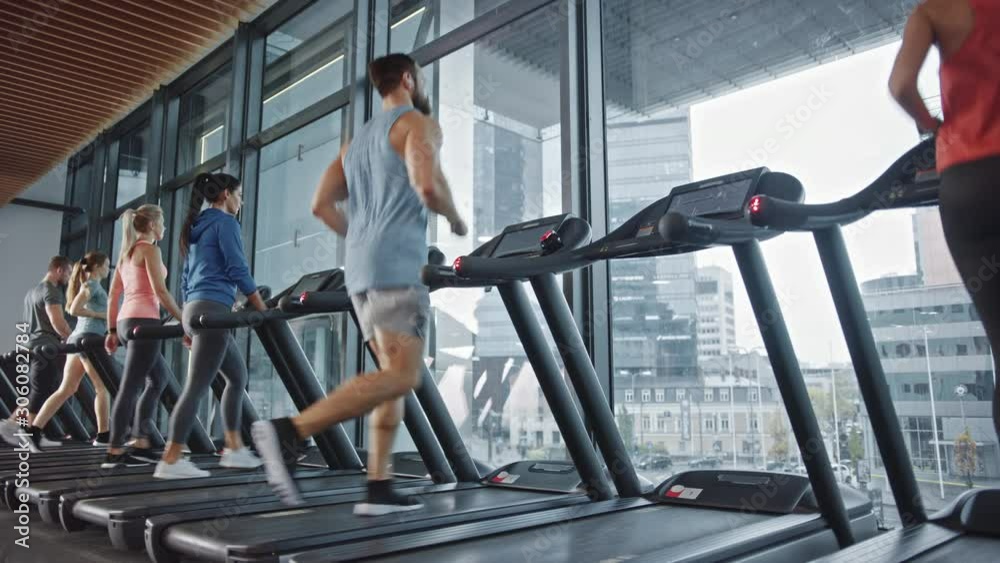 Muscular Male Athlete Puts on Wireless Headphones, Listens to a Podcast or Sport Music Playlist while Running on a Treadmill. In Background Fit Beautiful Women Training. Elevating Side View