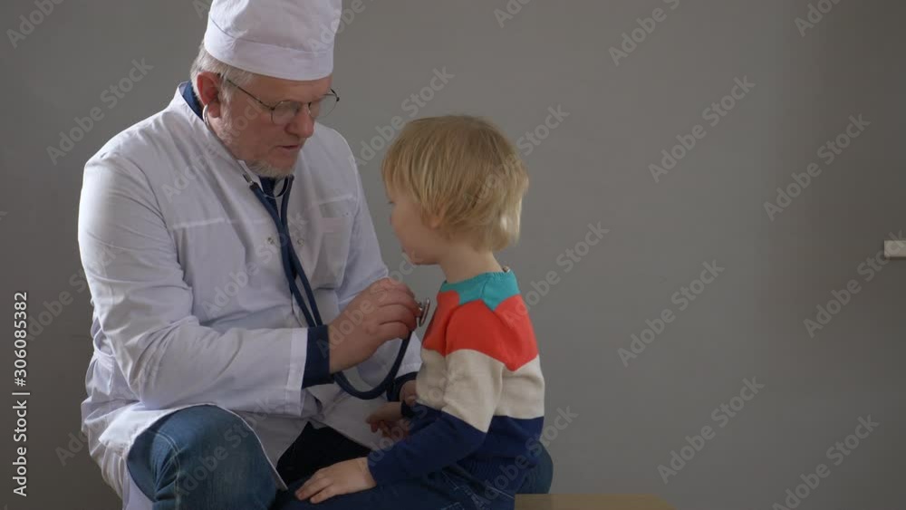 elderly doctor with gray hair and glasses in white robe and cap examines cute little blonde boy with stethoscope. doctor examines throat and listens to lungs. Child trusts medical man elderly doctor with gray hair and glasses in white robe and cap examines cute little blonde boy with stethoscope. doctor examines throat and listens to lungs. Child trusts medical man