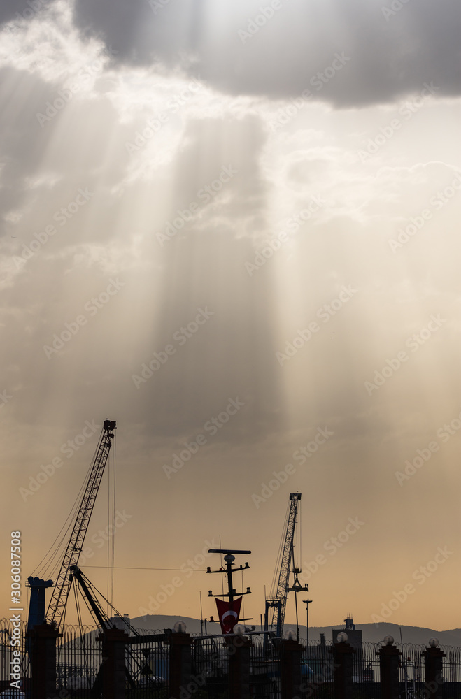 Dark silhouettes of industrial port cranes. Sun lights descending through the clouds. Alsancak coast, Izmir, Turkey.