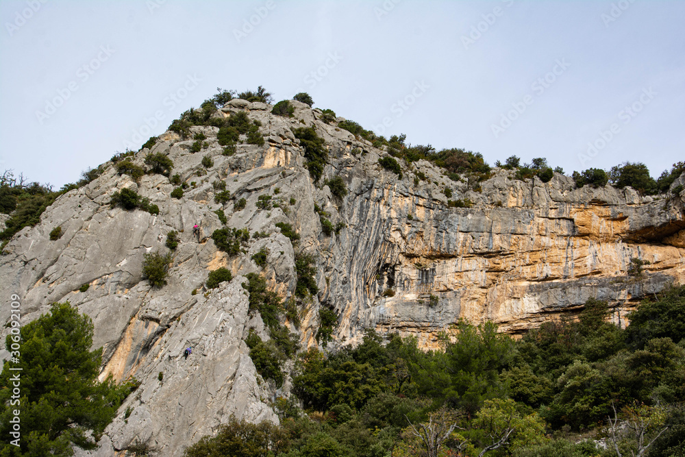 Climbers on a cliff in the south of France. Via Ferrata. Bushes, trees, a blue sky, grey and ocher colored rocks.