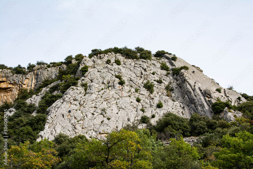 Several climbers climb up and down a cliff.  Via Ferrata in the south of France. Bushes, trees, a blue sky, grey and ocher colored rocks.