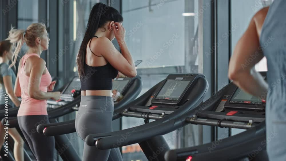 Athletic Sports Woman in Gym Puts on Wireless Headphones, Turns on Sport Music Playlist with Smartphone and Starts Running on Treadmill. In Background Fit Athletes Training. Slow Motion Side View Athletic Sports Woman in Gym Puts on Wireless Headphones, Turns on Sport Music Playlist with Smartphone and Starts Running on Treadmill. In Background Fit Athletes Training. Slow Motion Side View