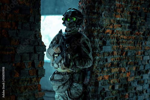 Man in uniform with machine gun and turned on night vision device stand between two brick walls. Airsoft soldier with green light on face in night photo