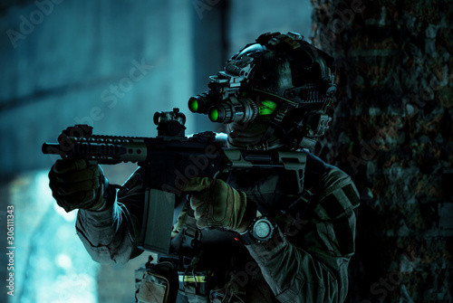 Man in uniform with machine gun and turned on night vision device inside broken building. Airsoft soldier with green light on face in night building