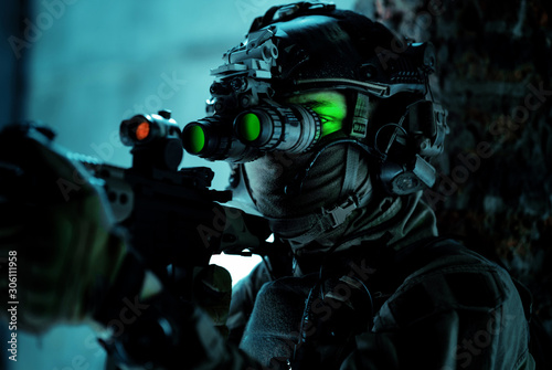 Man in uniform with machine gun and turned on night vision device beside brick wall. Closeup airsoft soldier with green light on face in night