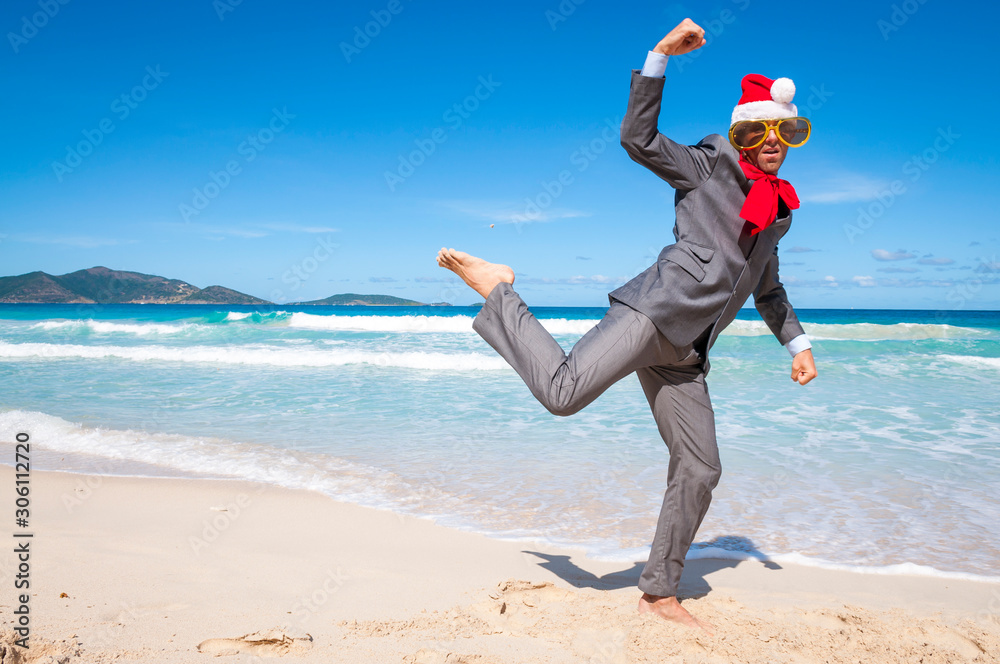 Dancing holiday businessman wearing Santa hat, sunglasses and big red Christmas bow celebrating barefoot on a tropical beach Dancing holiday businessman wearing Santa hat, sunglasses and big red Christmas bow celebrating barefoot on a tropical beach
