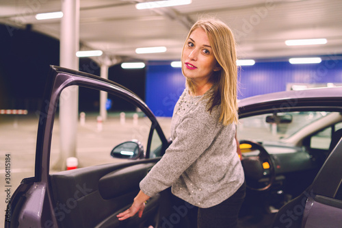 Young woman getting out of the parked car looking to the camera smiling on the parking in the city at night