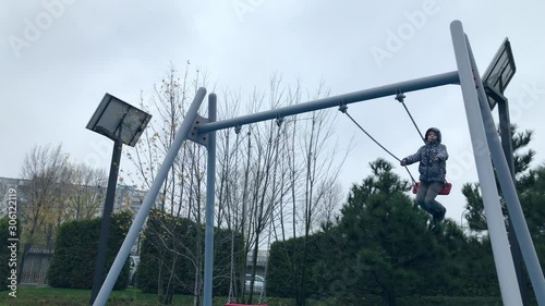Cheerful European boy swinging on a swing. A joyful child swings on a swing in the Playground.  Happy child swinging very high on a swing in the street. Fall season. Cloudy weather.   Low angle. photo