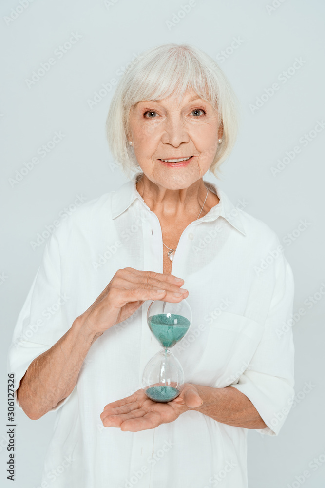 attractive and smiling woman looking at camera and holding hourglass isolated on grey attractive and smiling woman looking at camera and holding hourglass isolated on grey