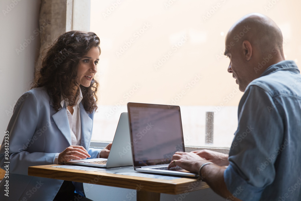 Two colleagues using laptops during break in cafe. Young woman and middle aged man sitting at table near window and working on computers. Coworkers concept Two colleagues using laptops during break in cafe. Young woman and middle aged man sitting at table near window and working on computers. Coworkers concept