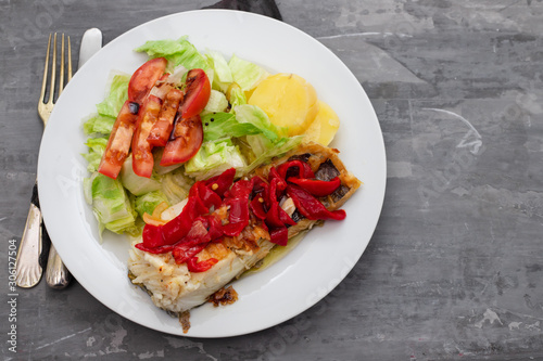 cod fish with red pepper, potato and salad on white plate on ceramic background