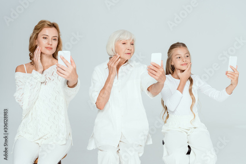 smiling granddaughter, mother and grandmother taking selfies and touching faces isolated on grey
