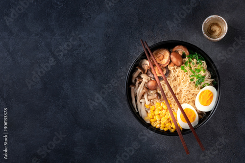 Ramen. Soba noodles with an egg, shiitake and enoki mushrooms, corn and scallions, shot from above on a dark background with chopsticks, sake, and a place for text