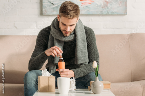 sick man sitting on sofa and opening bottle with cough syrup photo