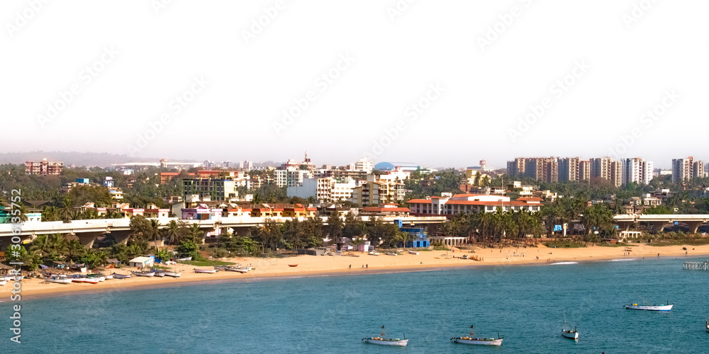 Beautiful Aerial view of Goan Beach from a cliff in India. This coastal area is well developed city center with buildings, flyover and greenery. Fishing Boats could be seen anchored in calm blue ocean