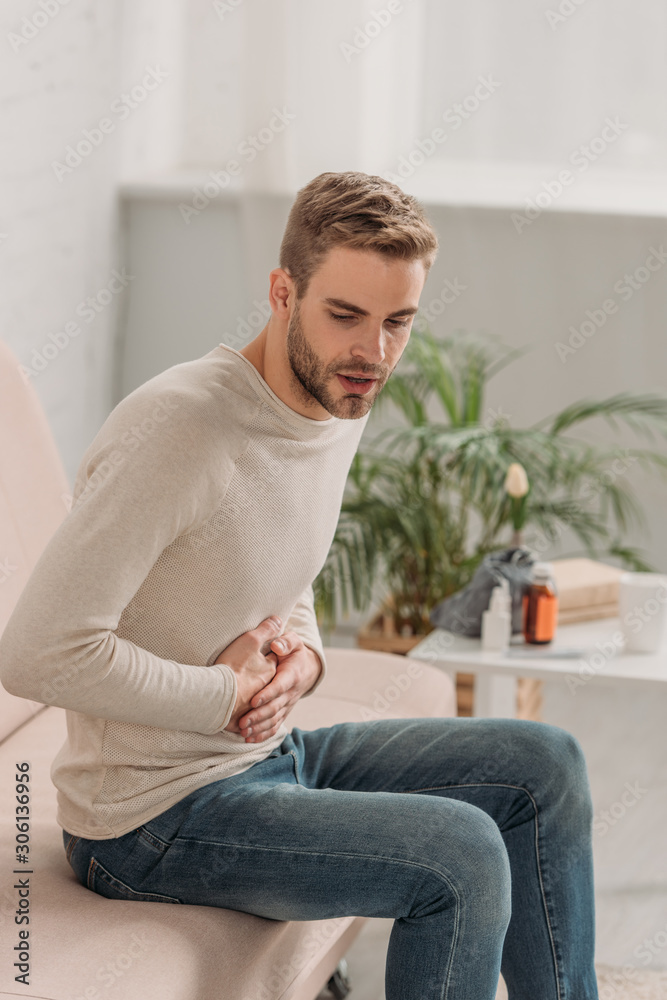 young man sitting on sofa and touching stomach while suffering from abdominal pain