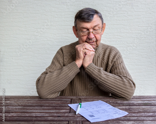 Old senior man looks with surprise on utility bills on wooden table. Planning month budget, calculating expen. Wrinkled palm of close up, copy space photo