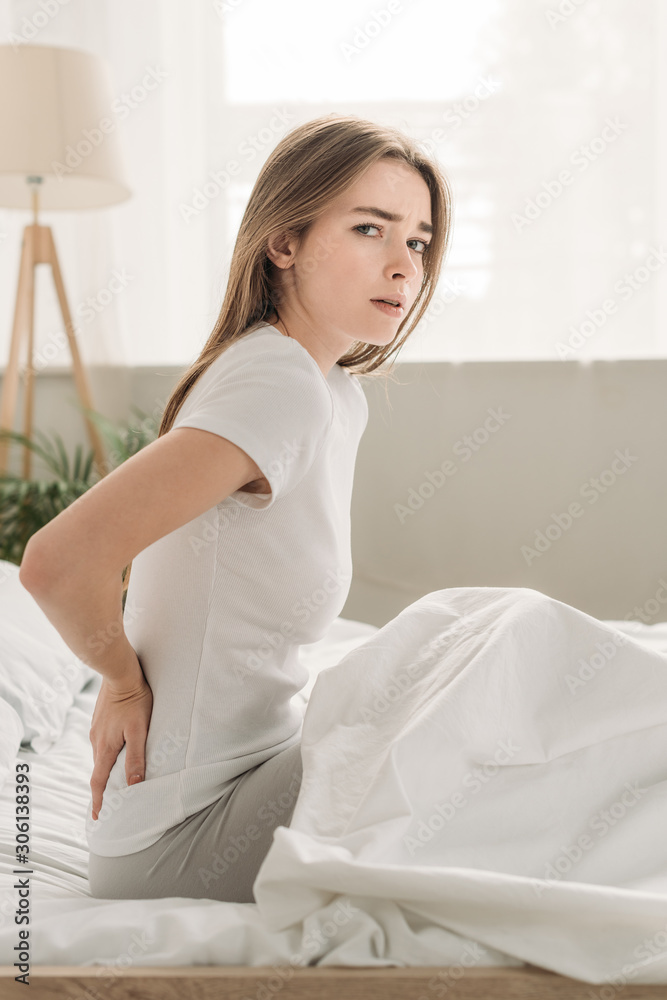 upset girl looking at camera while sitting in bed and suffering from pain in back upset girl looking at camera while sitting in bed and suffering from pain in back