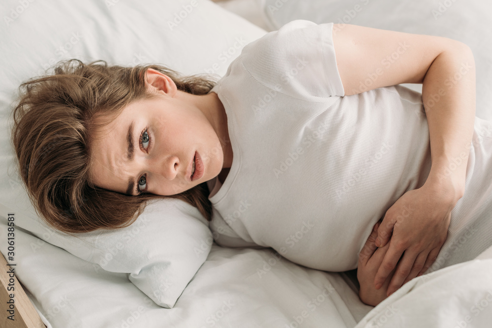 frowned girl looking at camera while lying in bed and suffering from abdominal pain frowned girl looking at camera while lying in bed and suffering from abdominal pain