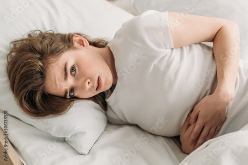 frowned girl looking at camera while lying in bed and suffering from abdominal pain