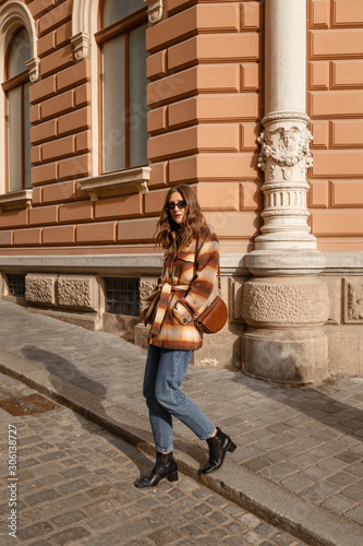 fashion outfit perfect for autumn fall winter. street style portrait of an attractive woman wearing plaid check jacket coat, denim jeans, black ankle boots and brown leather bag, crossing the street. 