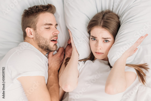 top view of woman plugging ears with pillow while lying in bed near snoring husband photo