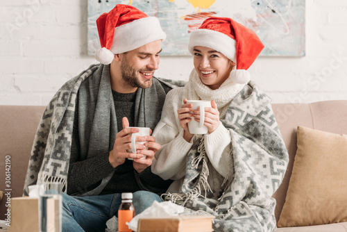 diseased, smiling couple in santa hats, covered with blanket, holding cups of warming drink while sitting on sofa
