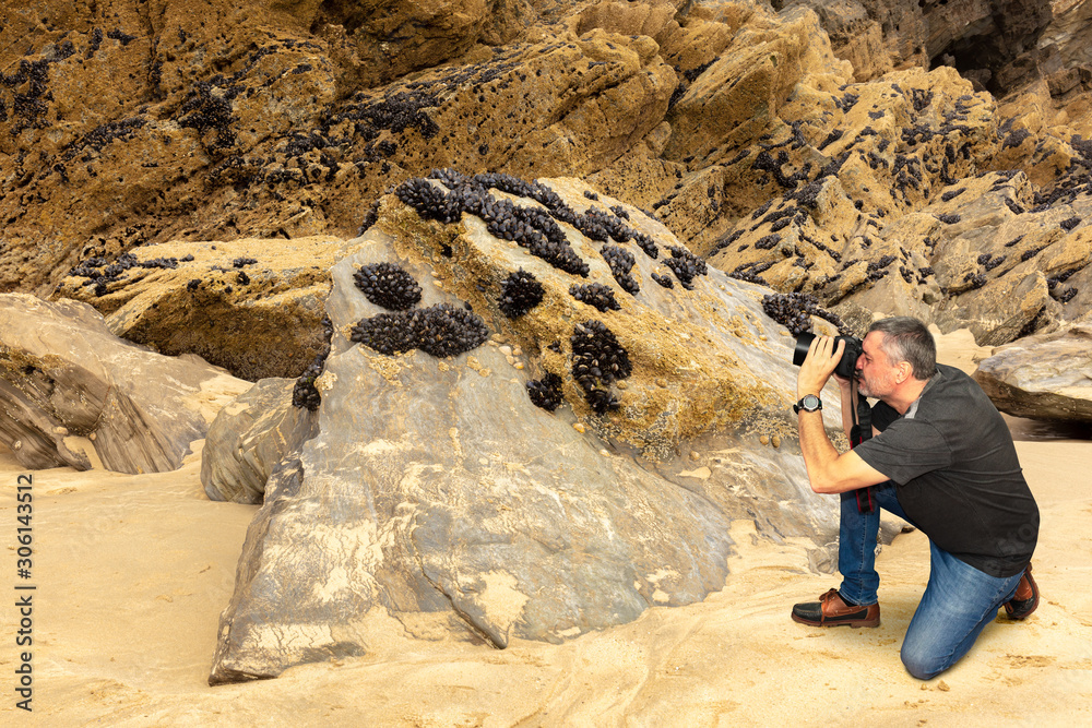 A photographer on the beach of Newquay in the English county of Cornwall. He photographs dark blue shells on a rock. The man kneels in the sand. A photographer on the beach of Newquay in the English county of Cornwall. He photographs dark blue shells on a rock. The man kneels in the sand.