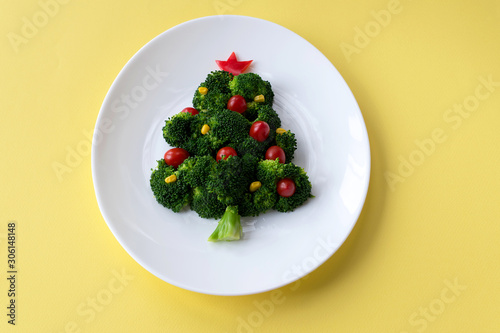 Christmas edible tree made from broccoli, tomato and corn on a white plate. Christmas card with gifts and dietary salad. Yellow background, copy space, flat lay.