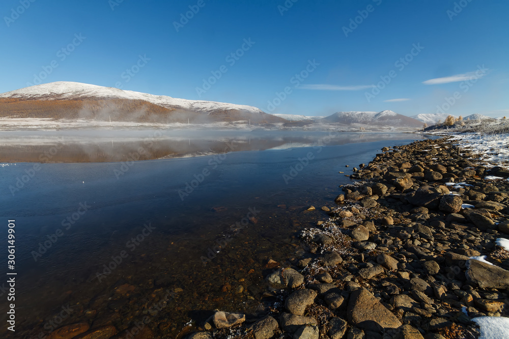 Obraz premium Mountain lake in Siberia in September with pebbles on the shore and reflection of snowy hills and blue sky