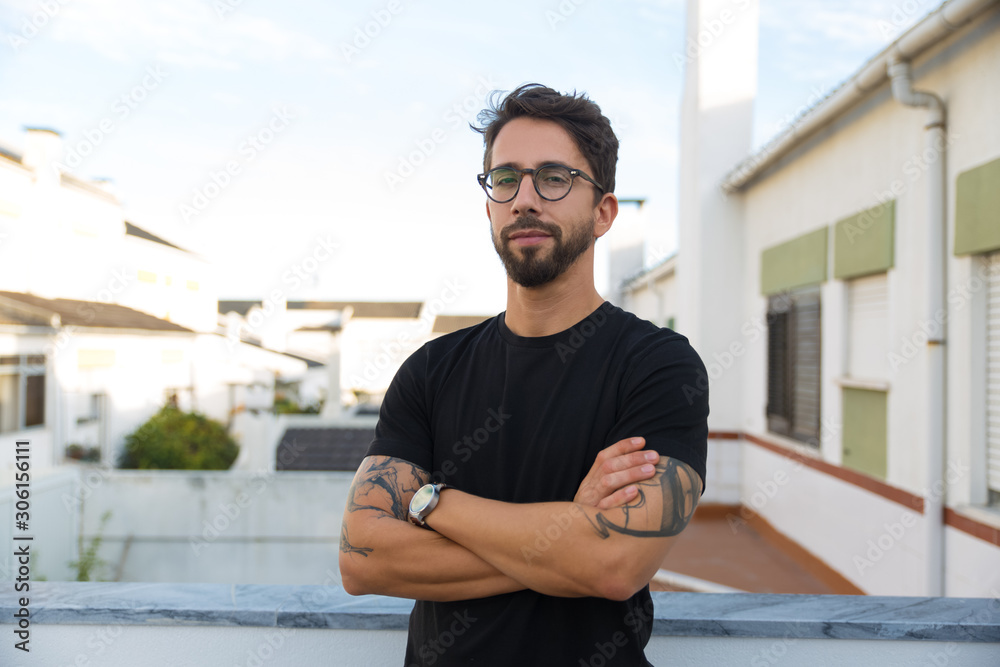 Confident stylish guy with tattoos posing on apartment balcony or terrace. Young man in glasses standing outside with arms crossed and looking at camera. Male portrait concept - obrazy, fototapety, plakaty 