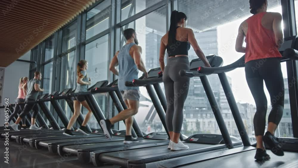 Fit Woman Walks onto Treadmill and Starts Running, Doing Her Fitness Exercise. Muscular Women and Men Actively Training in the Modern Gym. Sports People Workout in Fitness Club. Side View Slow Motion