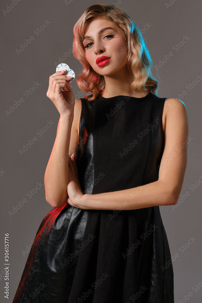 Blonde girl in black stylish dress holding two chips, posing against gray background. Gambling entertainment, poker, casino. Close-up. Blonde girl in black stylish dress holding two chips, posing against gray background. Gambling entertainment, poker, casino. Close-up.