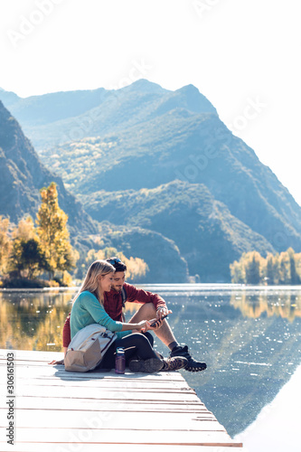 Two travel hikers using mobile phone while sitting in front of the lake in mountain.