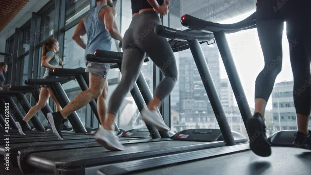 Fit Athletic Woman Walks onto Treadmill and Starts Running, Doing Her Fitness Exercise. Muscular Women and Men Actively Training in the Modern Gym. Low Ground Angle with Side View in Slow Motion