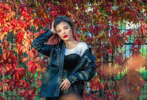 Portrait of beautiful stylish young woman with bright make-up, wearing black leather jacket, standing in front of bright colorful autumn foliage. 