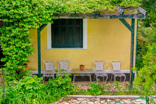 View of yellow house wall and terrace in the garden – white chairs, table, blooming flowers, green bushes and grass.