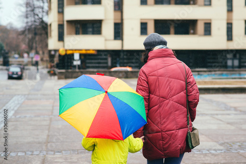 Mother with kid on a walk at city during rainy weather. Outfit, urban style. Colorful rainbow umbrella
