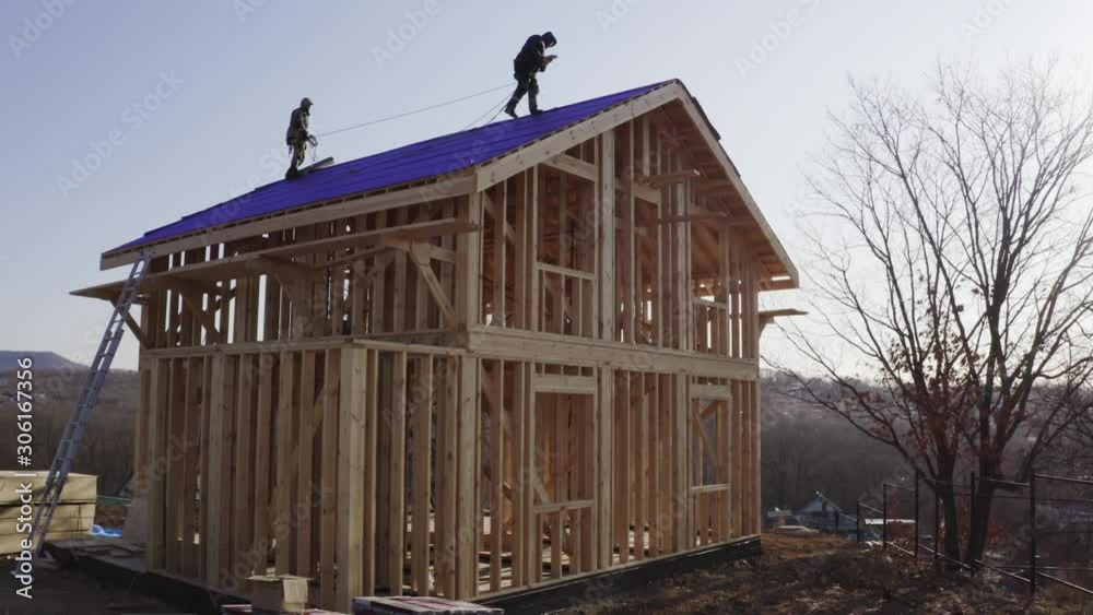 Two builders are working on the top of the roof covered with with insulation material. Aerial view of wooden frame house under construction Two builders are working on the top of the roof covered with with insulation material. Aerial view of wooden frame house under construction